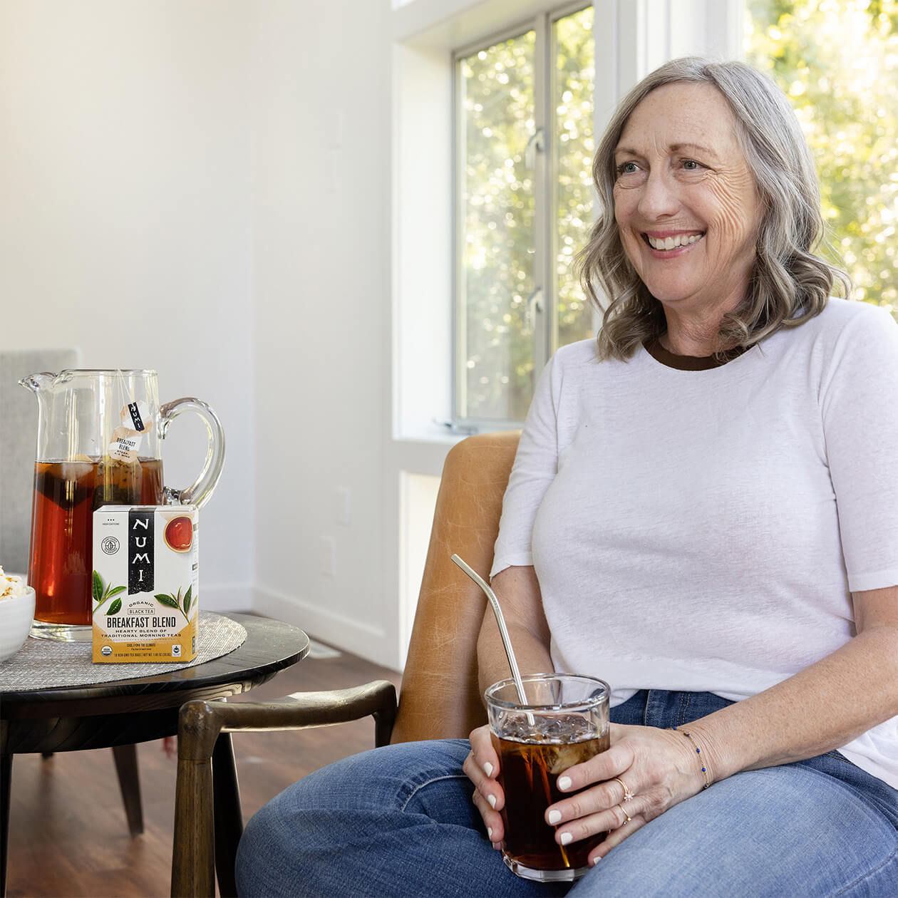 A middle aged woman in a tee shirt drinks iced Numi Breakfast Blend tea in her living room