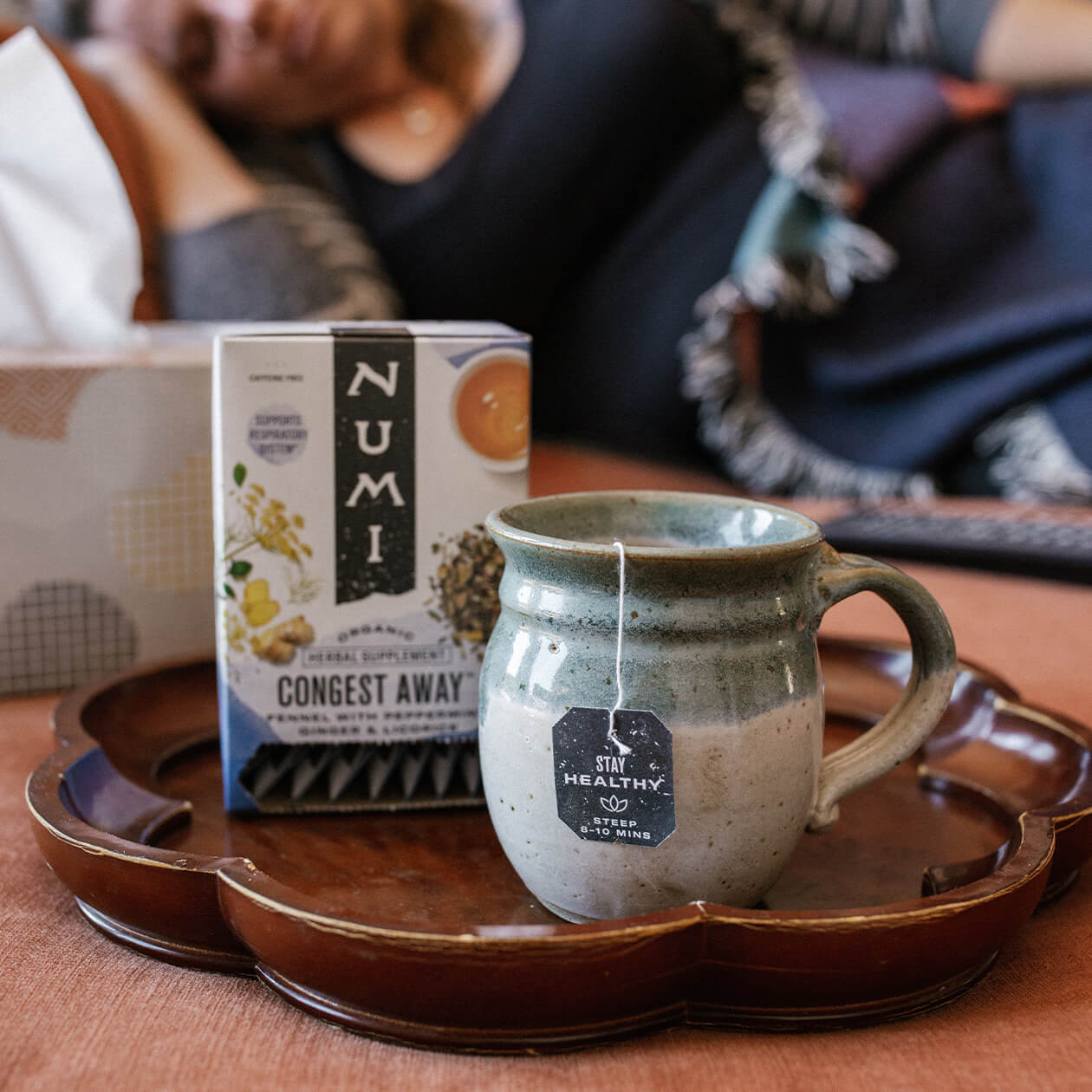 Numi's Congest Away tea brewing on a tray on a couch in front of a woman resting