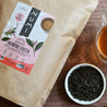 A bag of loose leaf Jasmine Green Tea sits on a wood table next to a mug of brewed tea